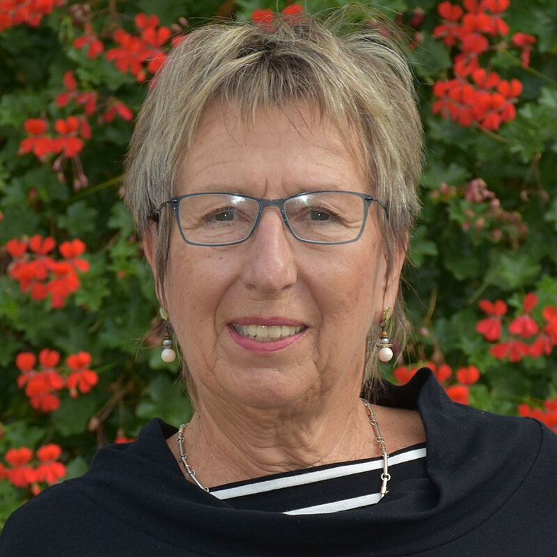 Leni Eggert Ältere Frau mit kurzen Haaren und Brille steht vor einer weißen Wand mit einem großen Blumenkasten voller roter Blumen. Sie trägt einen schwarzen Pullover mit gestreiften Ärmeln.