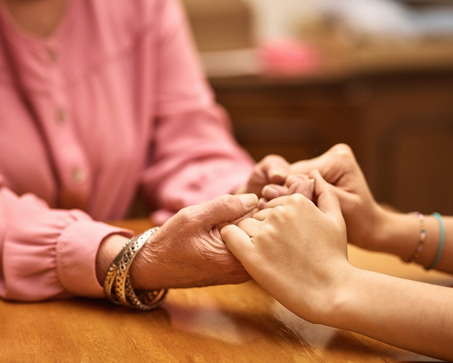 Hospizarbeit Hände halten Zwei Personen halten sich an den Händen, eine ältere mit Armbändern und eine jüngere, beide sitzen an einem Holztisch.