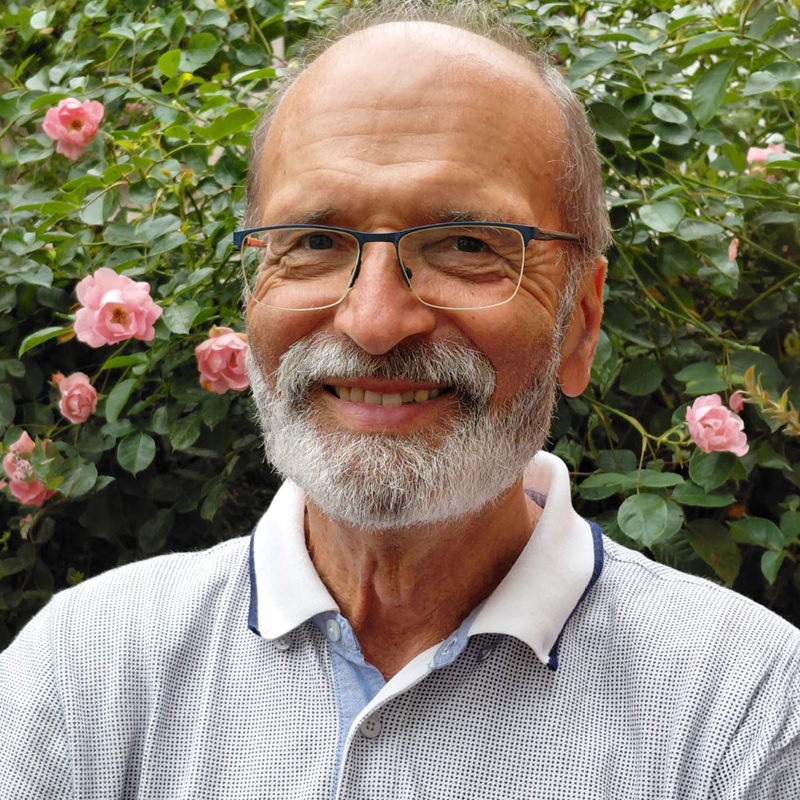 Erwin Fahr Ein älterer Mann mit Brille und grauem Bart lächelt vor einem Hintergrund mit grünen Blättern und rosa Rosen. Er trägt ein hellgraues Poloshirt.