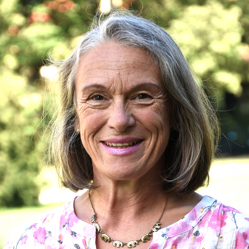 Michaela Gaus Porträt einer lächelnden älteren Frau mit grauem Haar, hellem Oberteil mit buntem Muster und einer Kette, aufgenommen im Freien vor verschwommenem Grün.