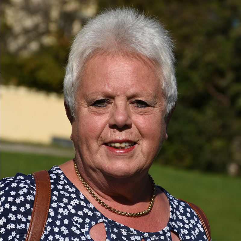 Edeltraud Wenzel Porträt einer älteren Frau mit kurzen weißen Haaren, die bei Sonnenschein im Freien steht und eine blau-weiße Bluse mit Blumenmuster sowie eine goldene Halskette trägt.